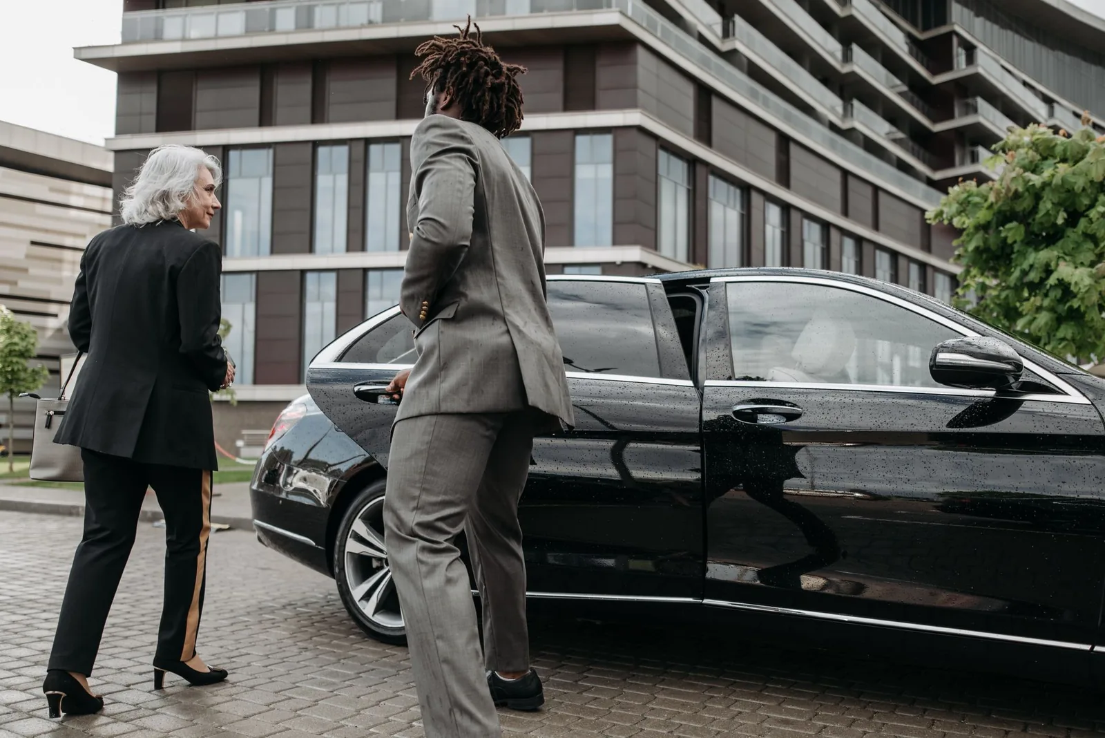 Chauffeur greeting two corporate passengers beside a black sedan outside an office building